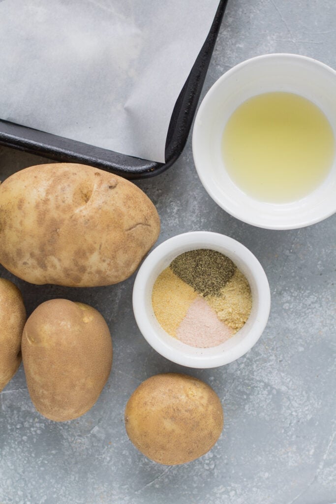 Ingredients for crispy roasted potatoes laid out on a grey background: olive oil, potatoes, salt and pepper, garlic powder (or minced garlic), onion powder