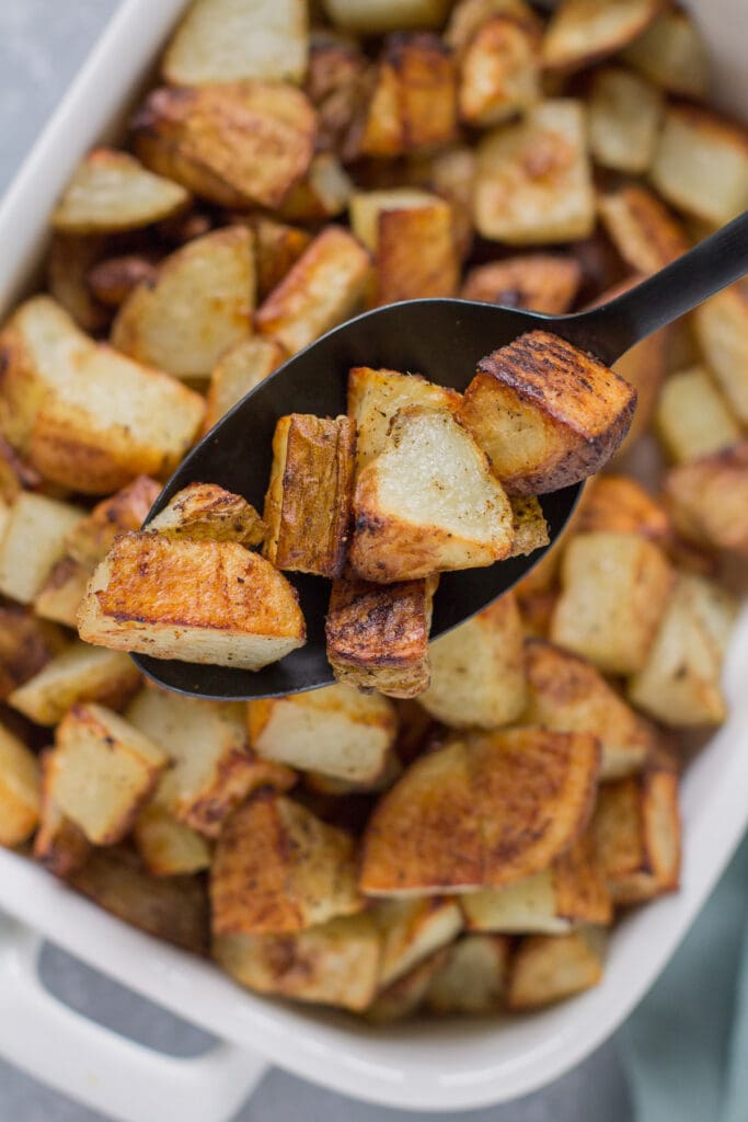 A spoon lifting up a scoop of crispy oven roasted potatoes out of a white baking dish with more seasoned potatoes in the background
