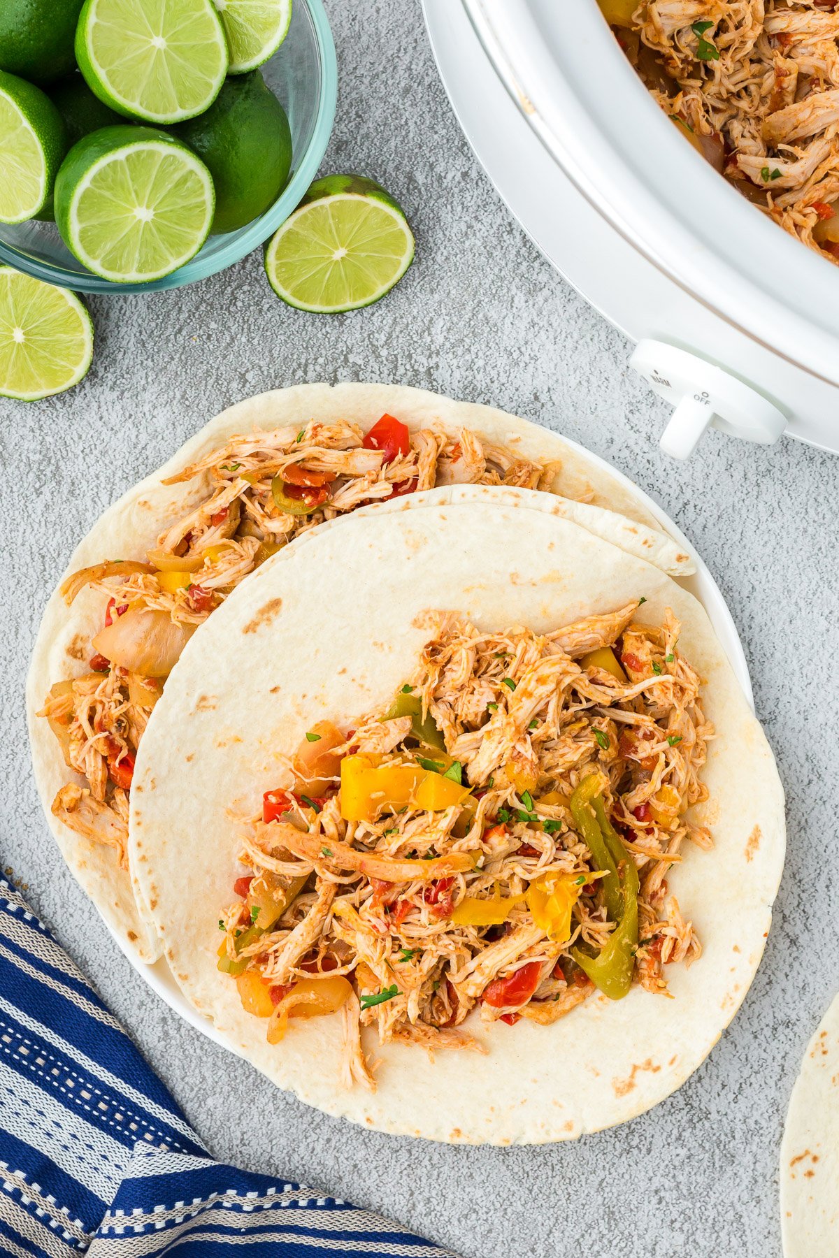 An overhead shot of two tortilla with crock pot chicken fajitas on a grey background with limes and a slow cooker on the side
