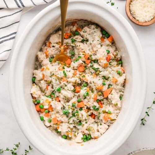 An overhead shot of crockpot chicken and rice in a white slow cooker base with a gold spoon in it with some herbs on the side on a white background.