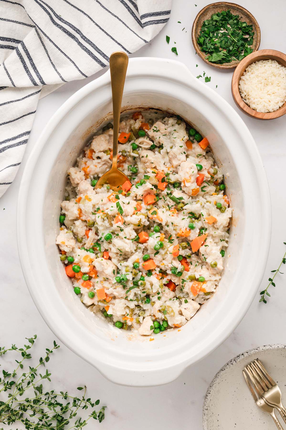 An overhead shot of crockpot chicken and rice in a white slow cooker base with a gold spoon in it with some herbs on the side on a white background.