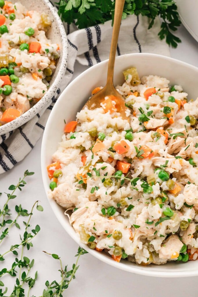 A white bowl with crockpot chicken and rice with a gold spoon in it on a white background with herbs on the side.