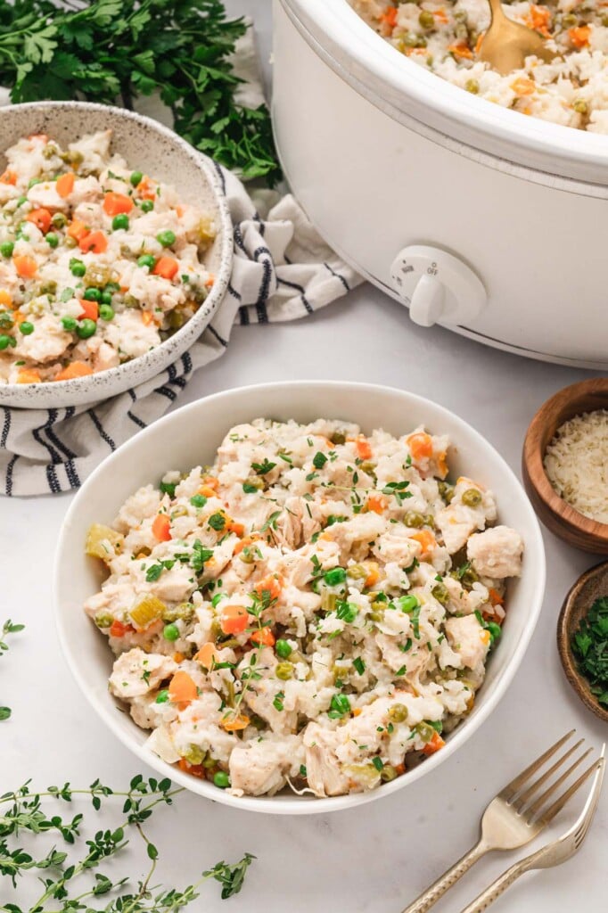 Two white bowls of crockpot chicken and rice with the slow cooker on the side on a white background with herbs on top.