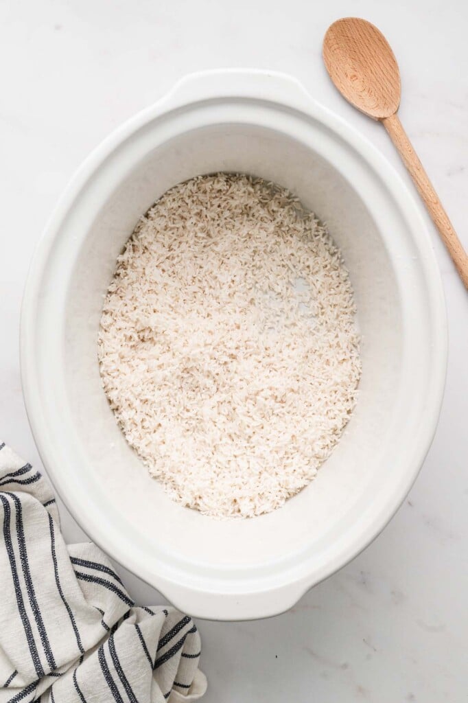 White rice laid in the bottom of a white crockpot base on a white counter with a wooden spoon on the side.