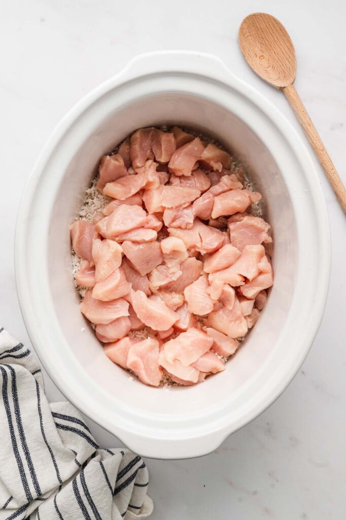 Raw chicken pieces on white rice in the bottom of a white crockpot base on a white counter with a wooden spoon on the side.