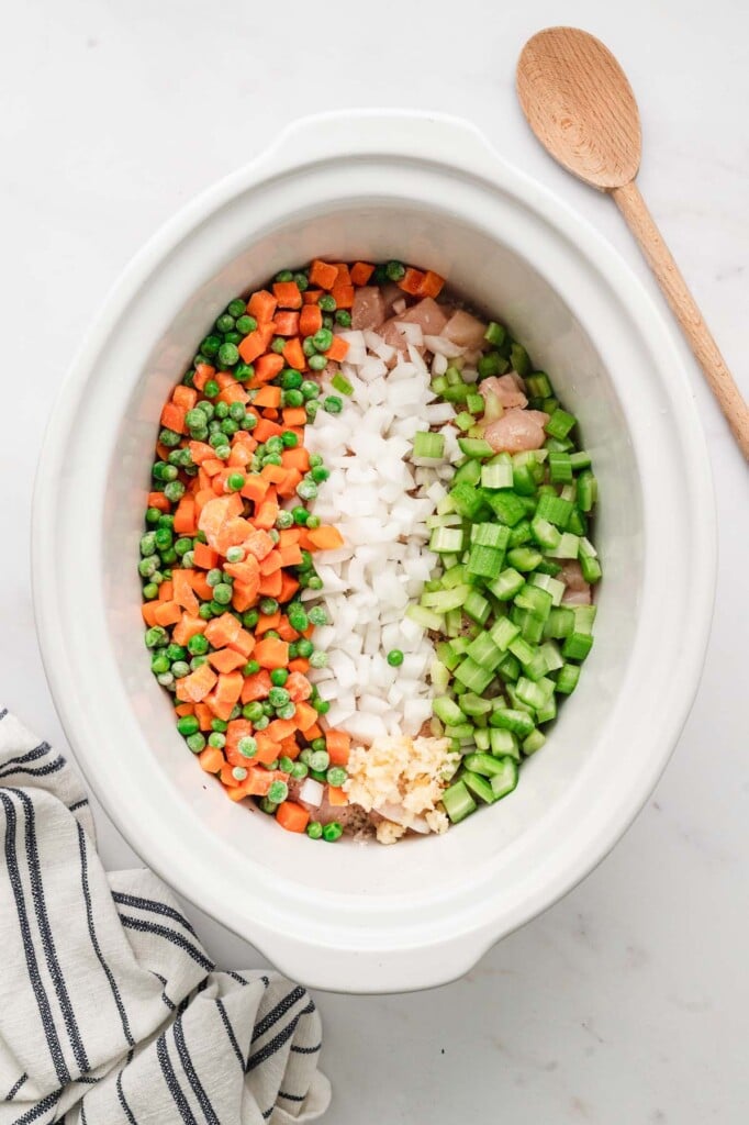 Onion, celery, and mixed carrots and peas on raw chicken pieces in a white crockpot base with a wooden spoon on the side on a white background. 