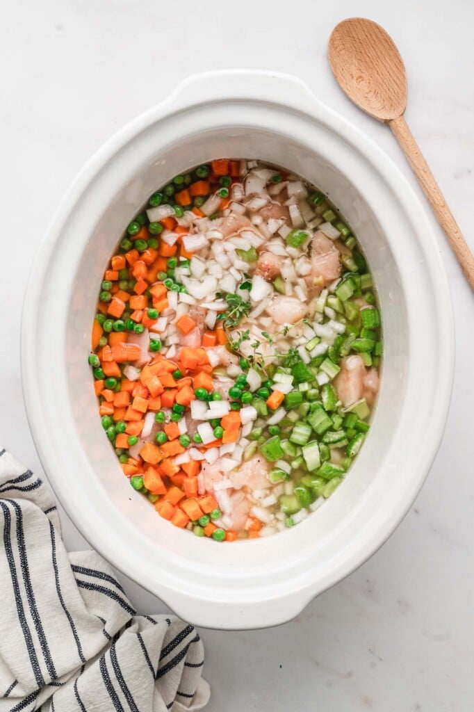 Crockpot Chicken and rice in a white slow cooker base after mixing together but before cooking on a white background with a wooden spoon on the side.