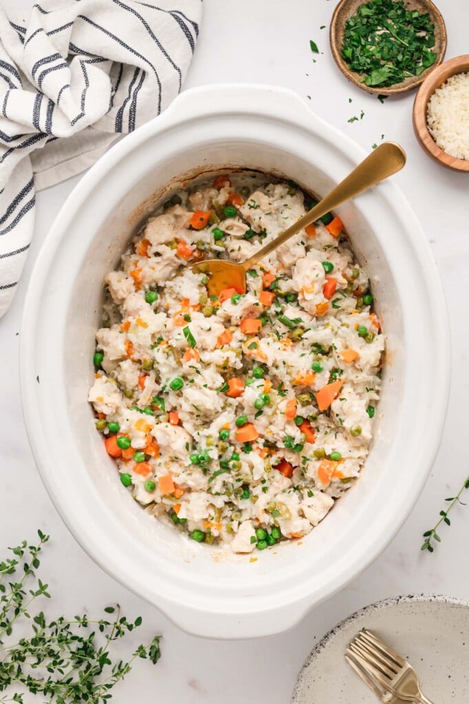 Cooked and mixed slow cooker chicken and rice in a white crockpot base with a gold spoon in it with herbs on the side.