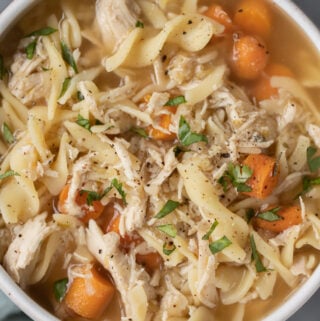 A close up of crockpot chicken noodle soup in a white bowl on a grey background