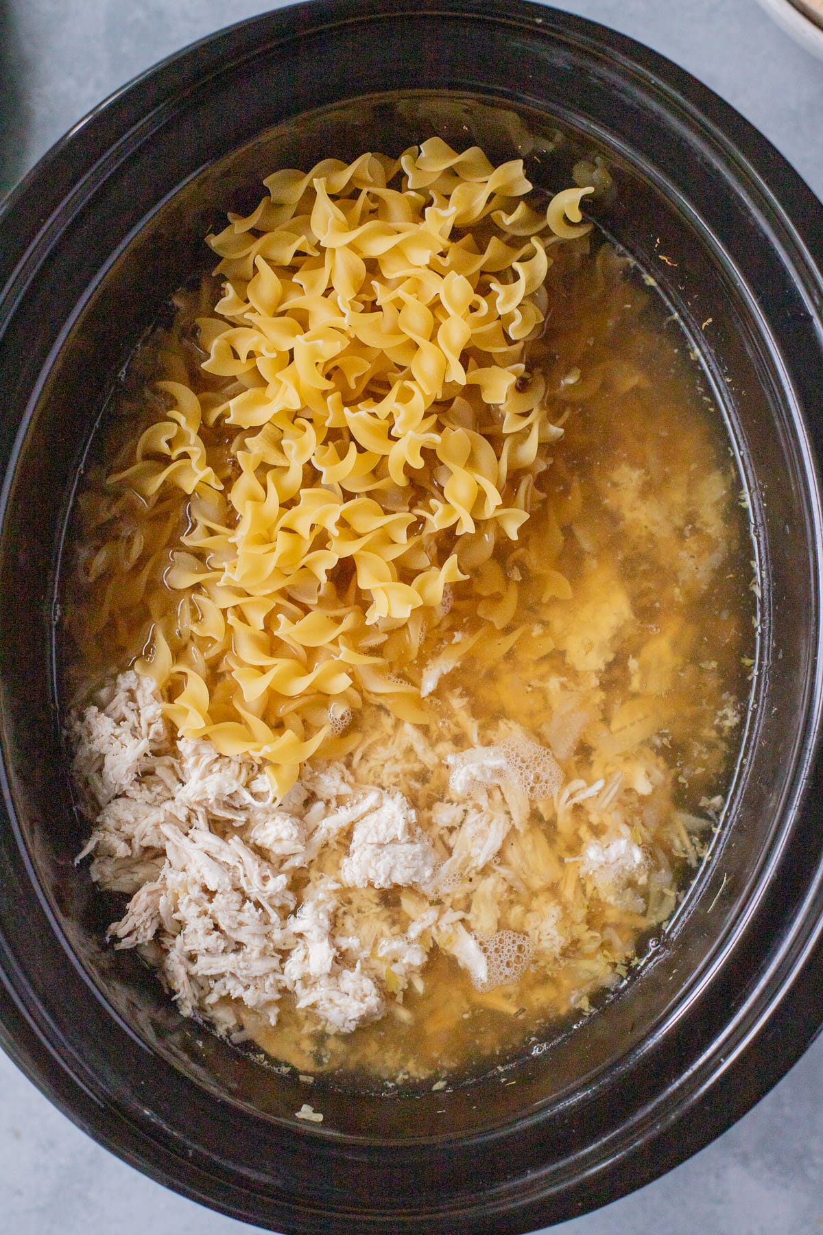 A close up of slow cooker chicken noodle soup with noodles being added in to cook in one pot