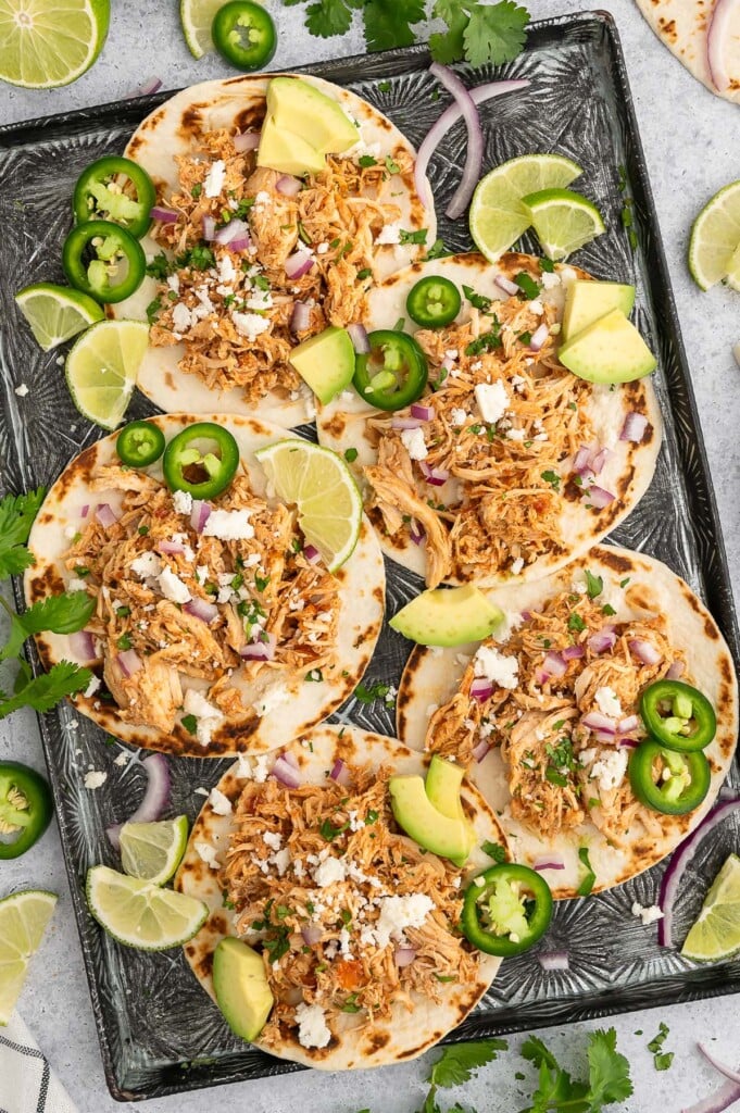 5 Crockpot Chicken Tacos on a baking sheet garnished with jalapeños, lime slices and avocado pieces.
