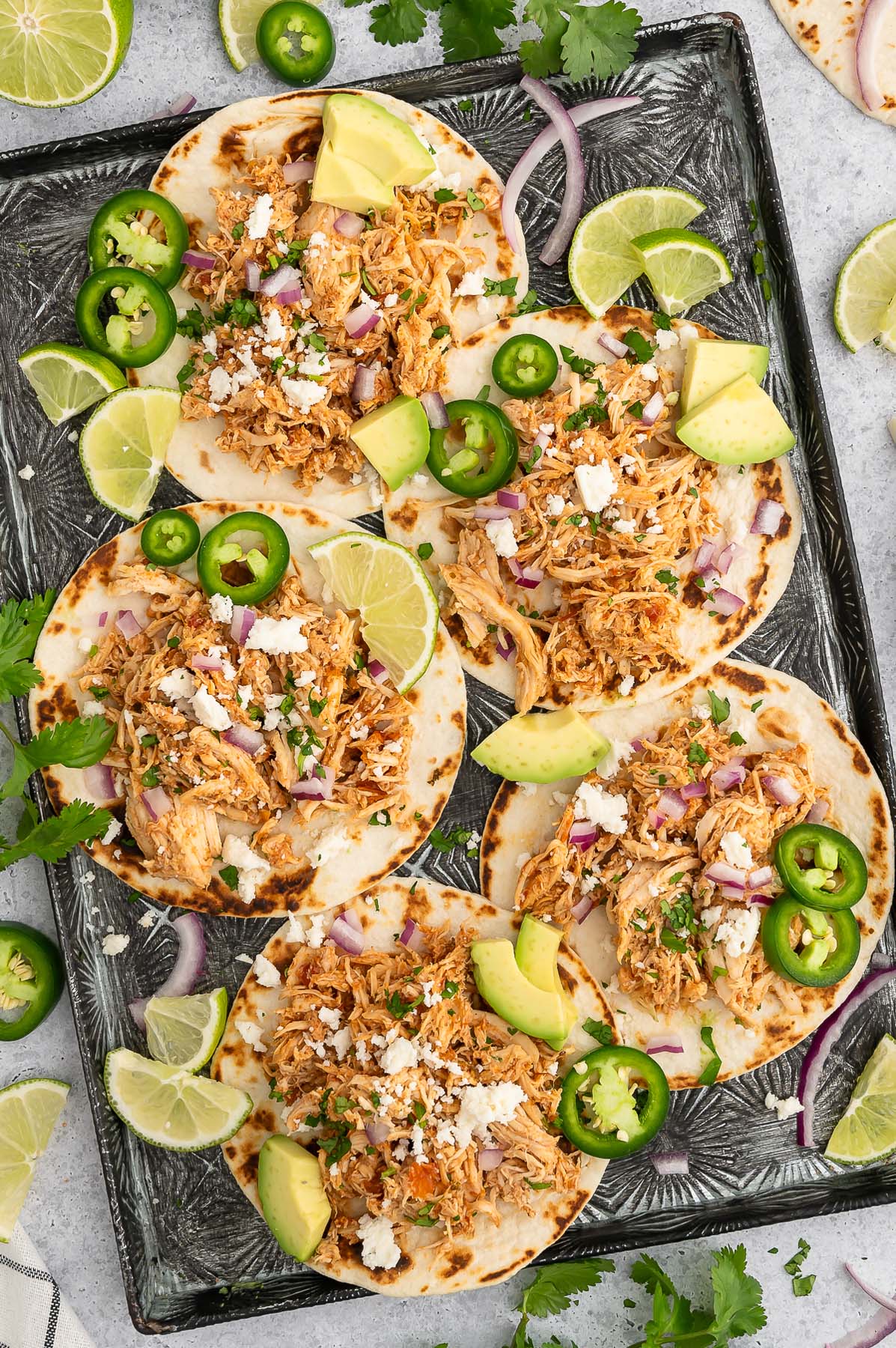 5 Crockpot Chicken Tacos on a baking sheet garnished with jalapeños, lime slices and avocado pieces.
