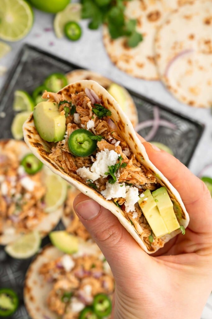A hand holding up a crockpot chicken taco garnished with avocado and jalapeños.