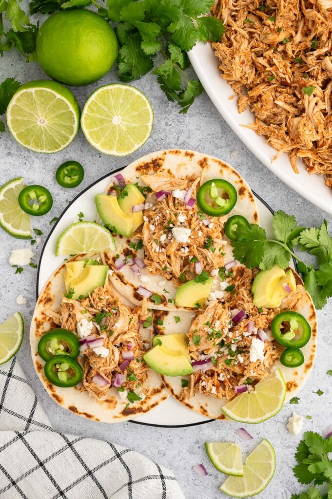 3 Crockpot Chicken Tacos on a white plate garnished with jalapeños, lime slices and avocado pieces.