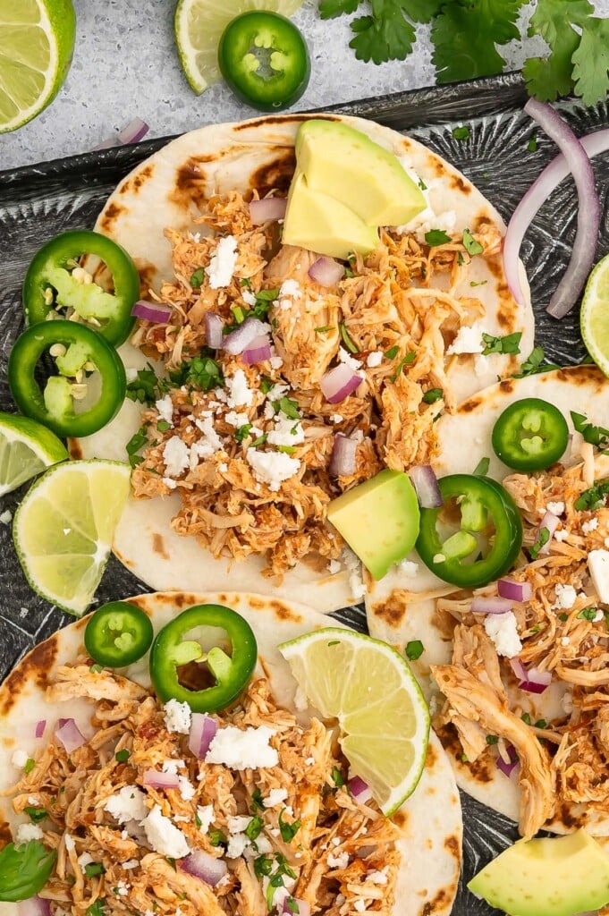 Crockpot Chicken Tacos on a baking sheet garnished with jalapeños, lime slices and avocado pieces.