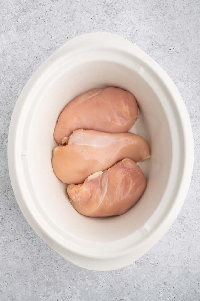 Three raw chicken breasts in a crockpot on a grey background.