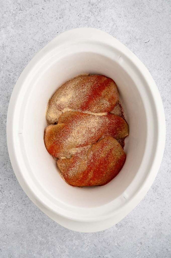 Three raw chicken breasts in a crockpot topped with taco seasoning.