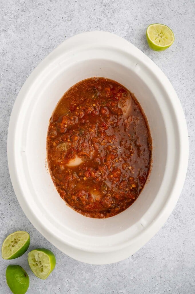 Uncooked crockpot chicken tacos in a white crockpot base with limes on the side.