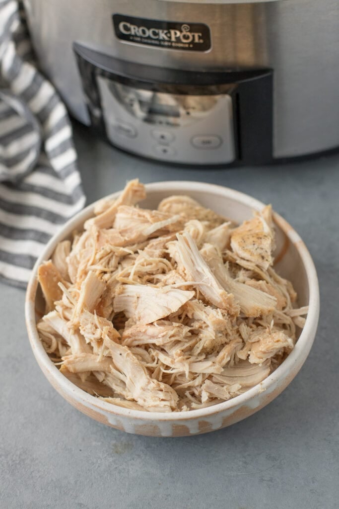 Crockpot shredded chicken in a white bowl on a grey background with crockpot in background