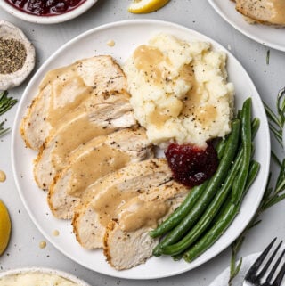 An overhead shot of crockpot turkey breast with gravy on top on a white plate with mashed potatoes and green beans