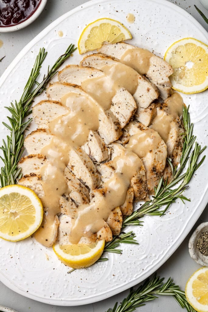 Crock pot Turkey breast with gravy on top on a white platter with herbs and lemons on the side