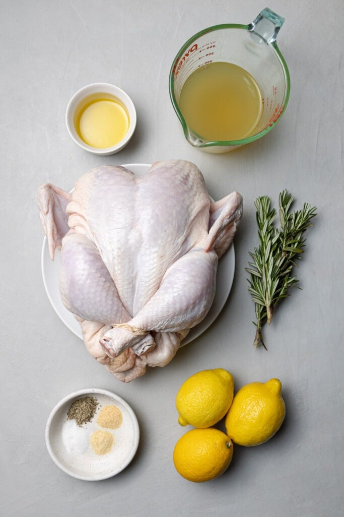 Ingredients for a crockpot whole chicken laid out in small bowls/dishes on a grey background: olive oil, rosemary, lemons, garlic powder, onion powder, salt, pepper, broth, whole chicken