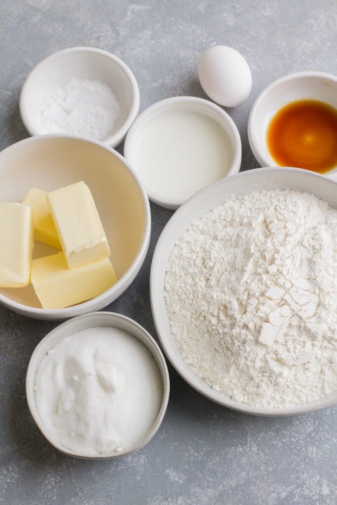 Cut out sugar cookie ingredients - butter, flour, sugar, milk, vanilla, an egg and baking powder in small white bowls