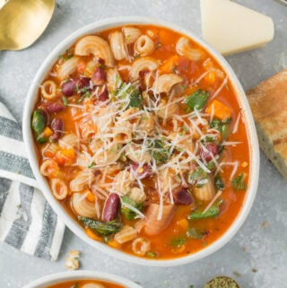 A close up of a bowl of easy minestrone soup with cheese, spoons and dried herbs off to the side.