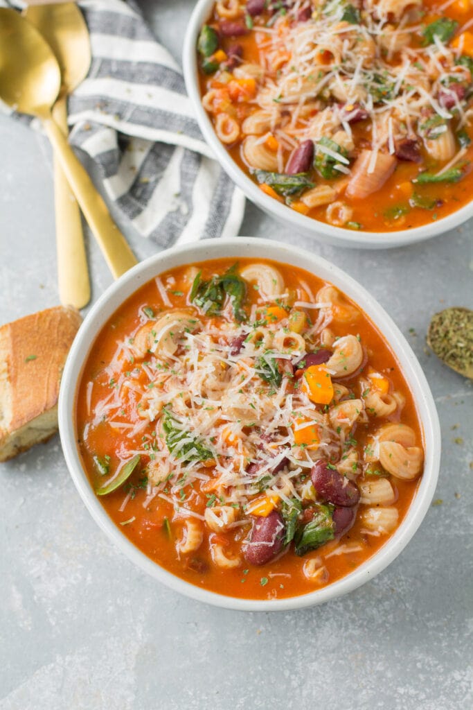 Two bowls of easy minestrone soup on a grey background with spoons and bread scattered off to the side.