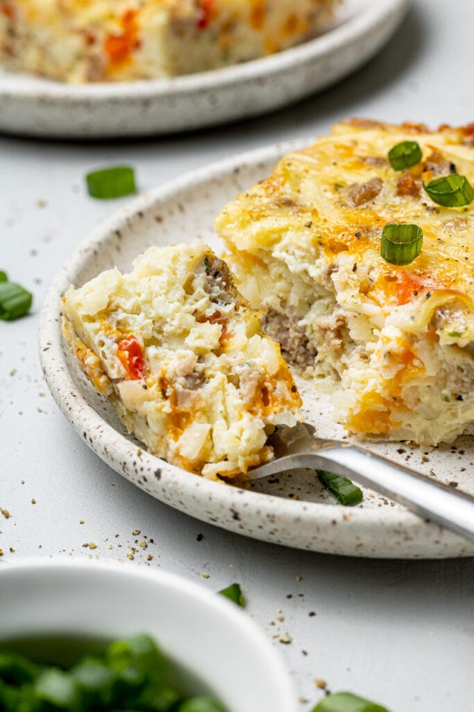 A piece of egg hash brown casserole on a plate with a fork with a bite of casserole on it