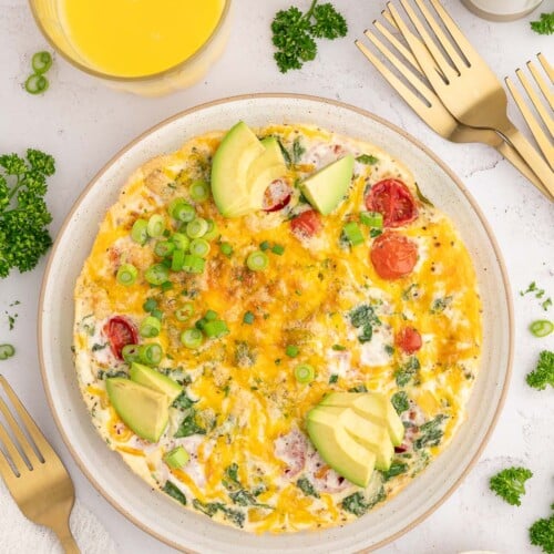 An Egg White Frittata on a plate garnished with sliced avocados and green onions with a glass of orange juice on the side.