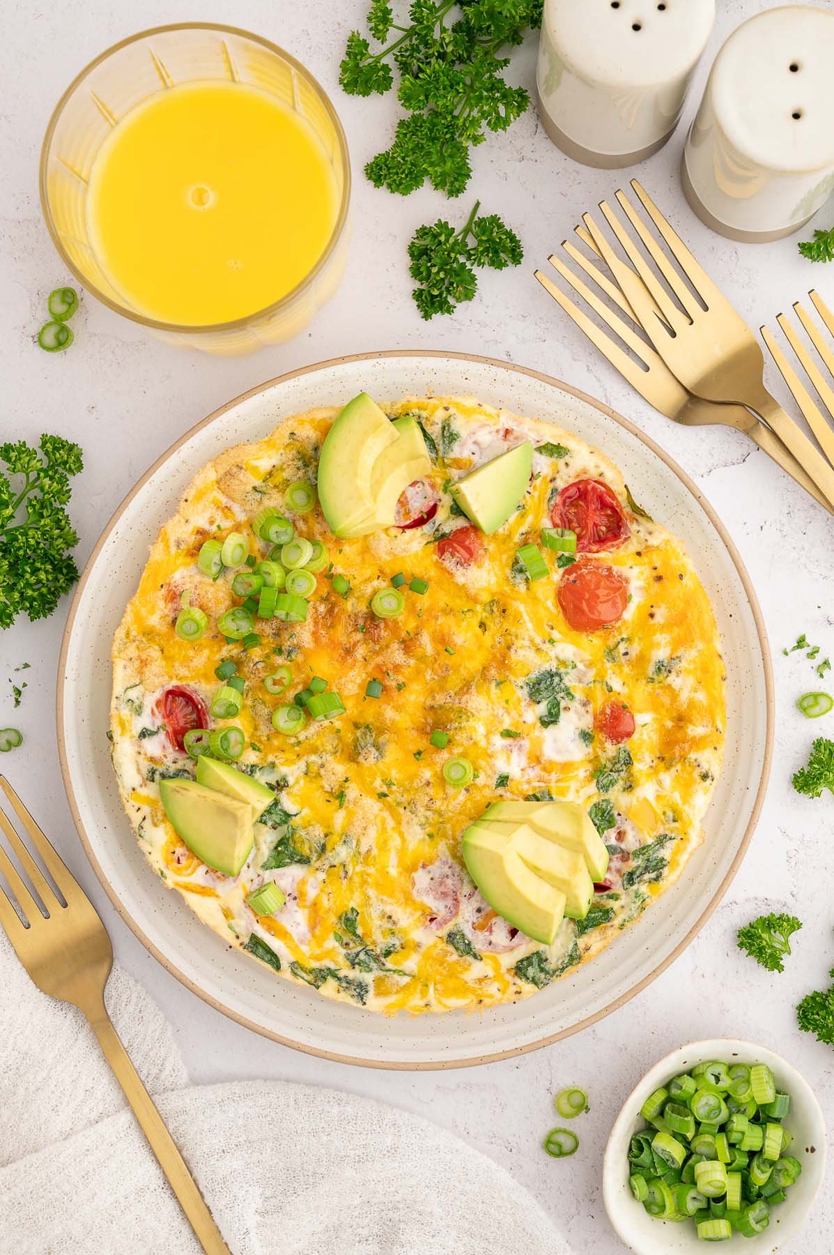 An Egg White Frittata on a plate garnished with sliced avocados and green onions with a glass of orange juice on the side.
