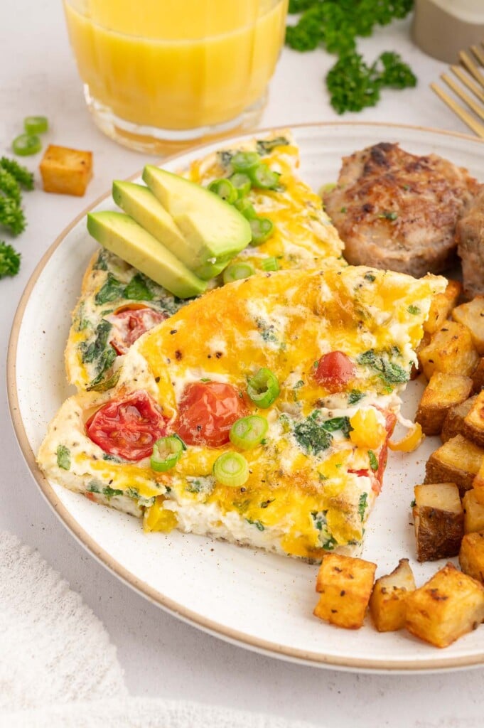 A slice of egg white frittata on a white plate garnished with green onions and avocado with chicken sausage and breakfast potatoes on the side.