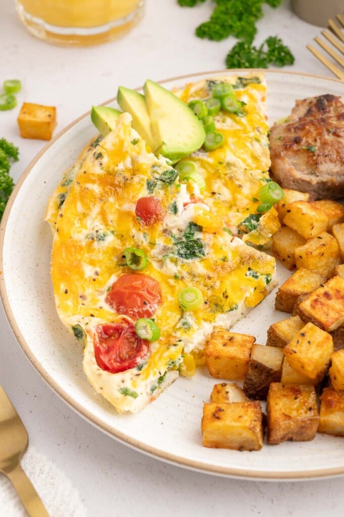 A slice of egg white frittata on a white plate garnished with green onions and sliced avocado with chicken sausage and breakfast potatoes on the side.