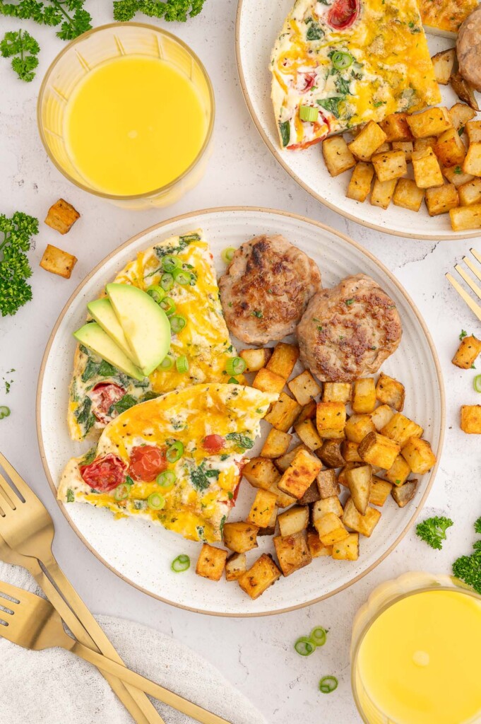 Two slices of egg white frittata on a white plate garnished with green onions and avocado with chicken sausage and breakfast potatoes on the side and a glass of orange juice on the side.