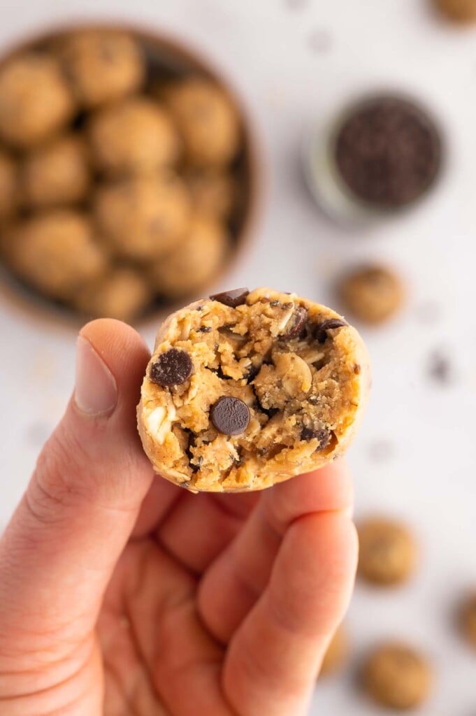 A peanut butter energy ball that has been bit in half with a hand holding it up with a stack of the balls in the background