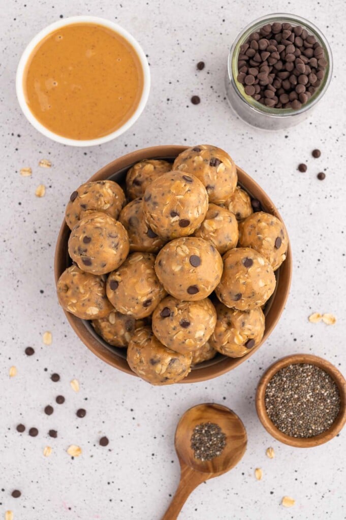 An overhead shot of peanut butter energy balls stacked on a brown plate with chia seeds, mini chocolate chips, and peanut butter on the side