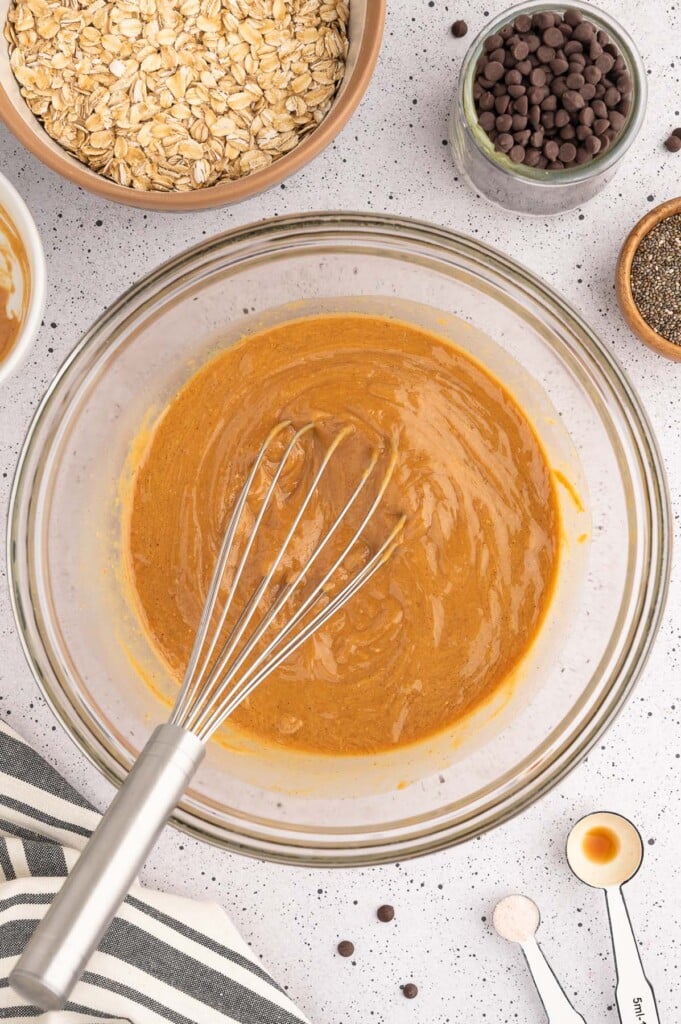 Peanut Butter, maple syrup, coconut oil, and vanilla extract being whisked together in a clear bowl with more ingredients for energy balls on the side