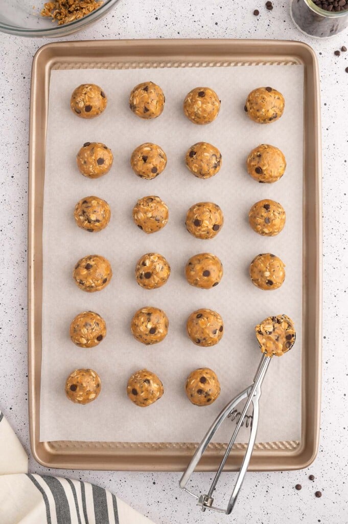 A cookie dough scoop and peanut butter energy balls on a parchment lined baking sheet on a white background