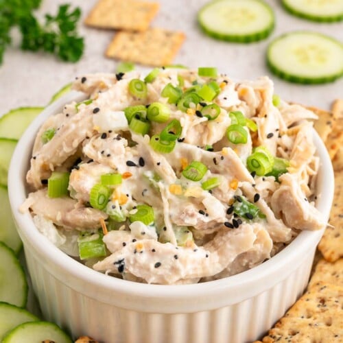Everything bagel chicken salad in a small white bowl topped with green onions with sliced cucumbers and crackers on the side.