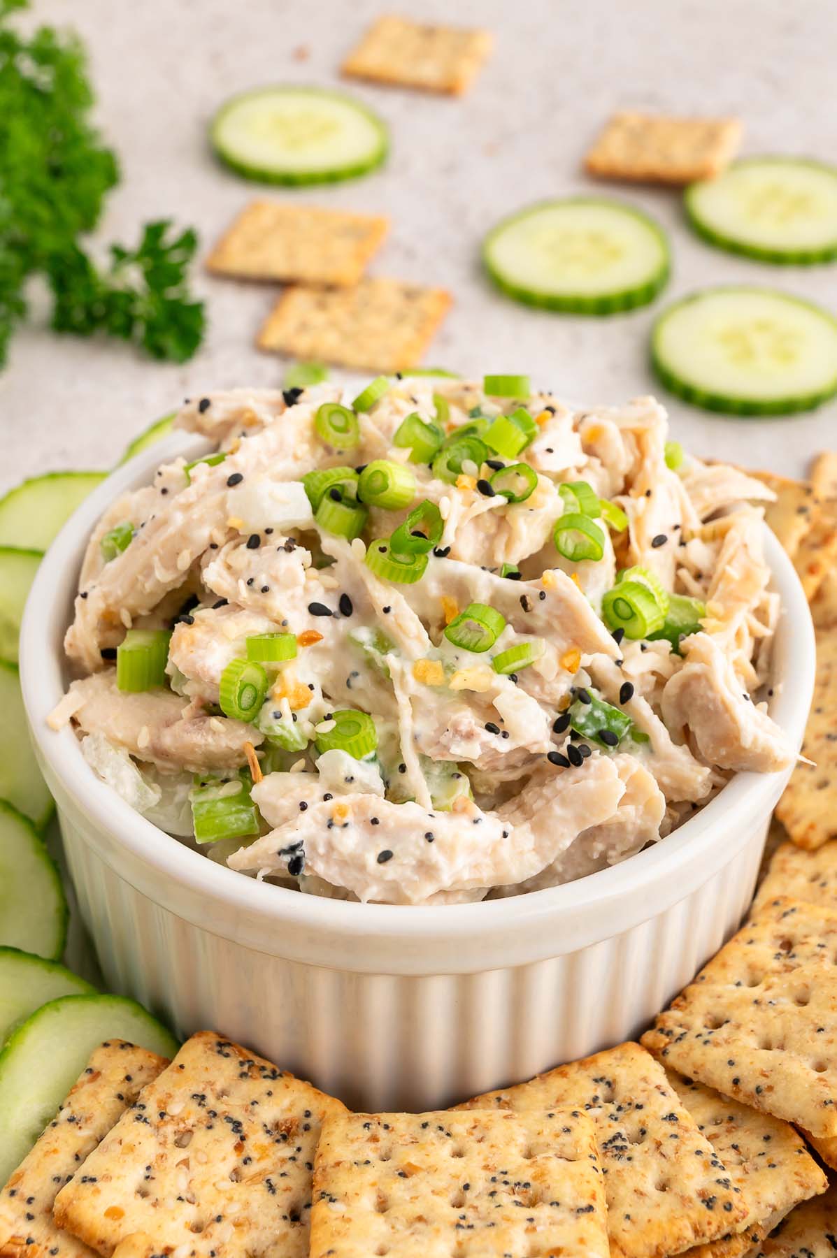 Everything bagel chicken salad in a small white bowl topped with green onions with sliced cucumbers and crackers on the side.