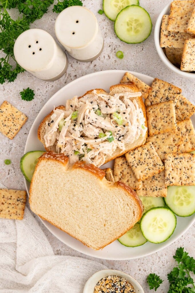 Everything Bagel Chicken Salad on a piece of bread with the top piece of bread for the sandwich on the side with crackers and sliced cucumbers also on the side.