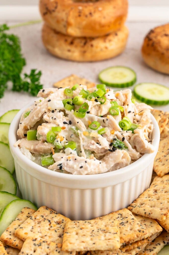 Everything bagel salad in a small white bowl topped with green onions with sliced cucumbers and crackers on the side.