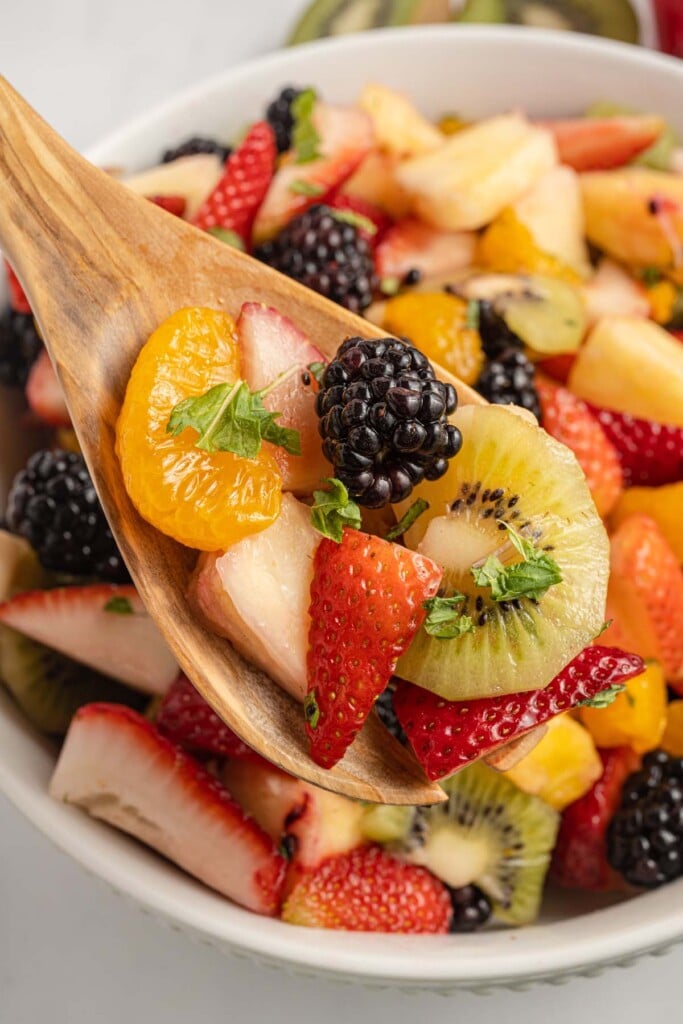 A wooden spoon taking a spoonful of fruit salad out of a large white bowl filled with the salad.