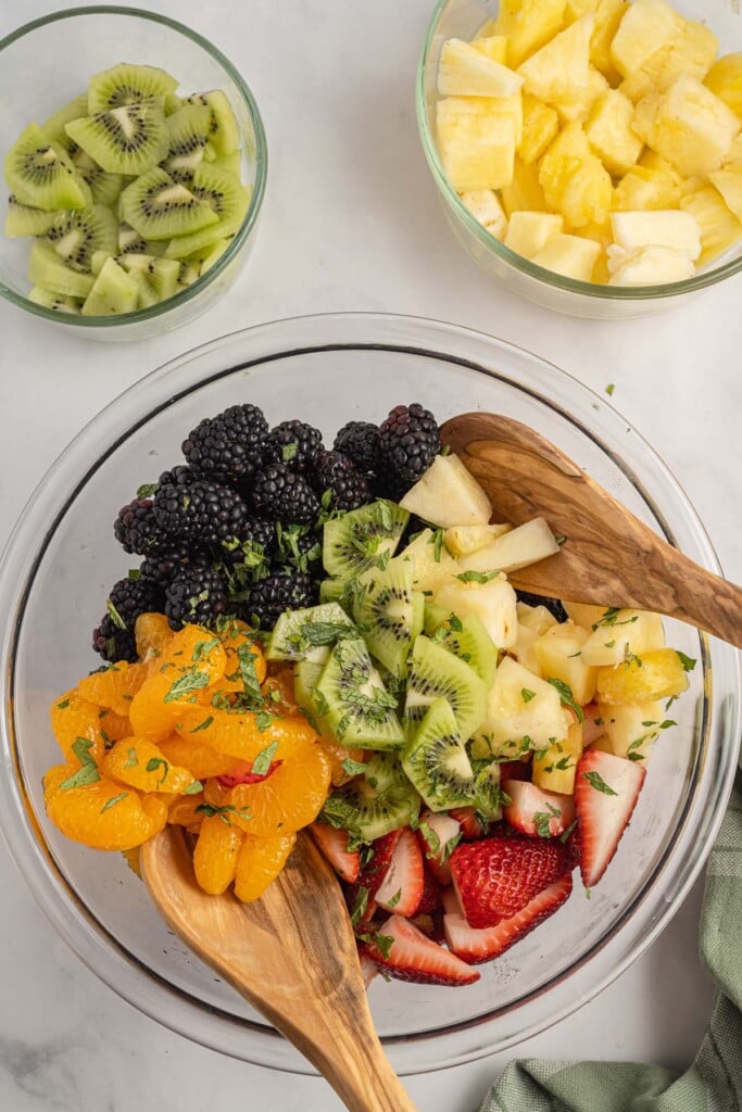 Fruit Salad being mixed with two wooden spoons in a clear bowl.