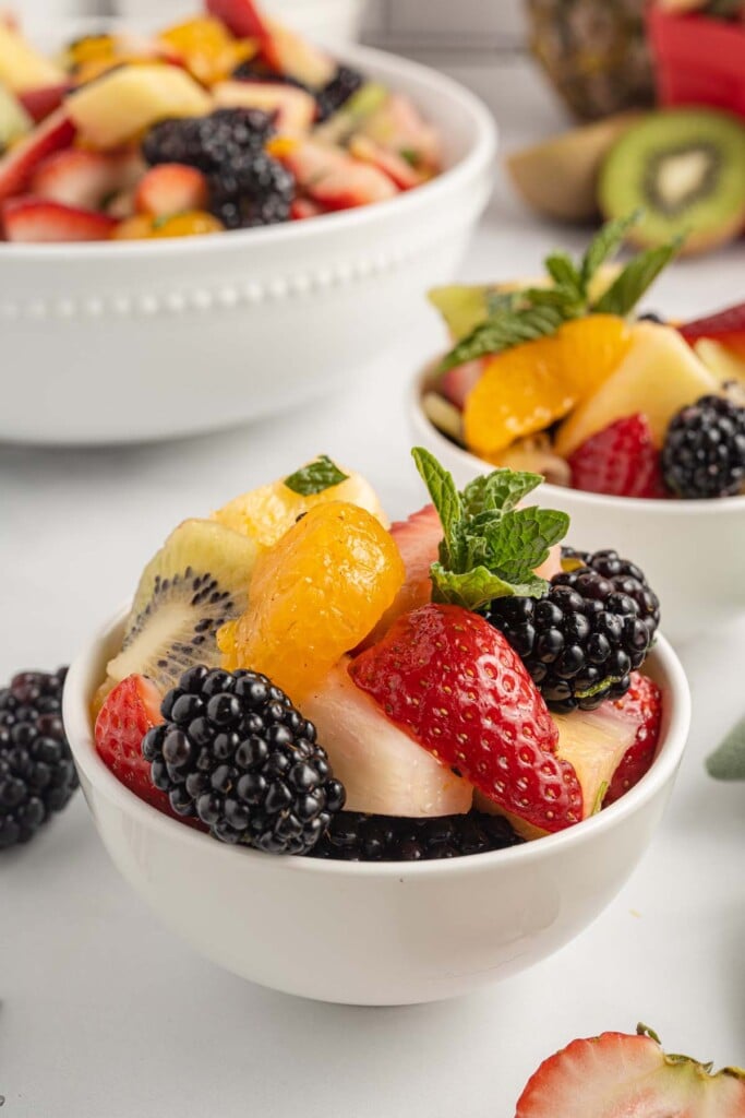 A small white bowl filled with fruit salad with mint garnishing the salad and a larger bowl of the fruit salad in the background.
