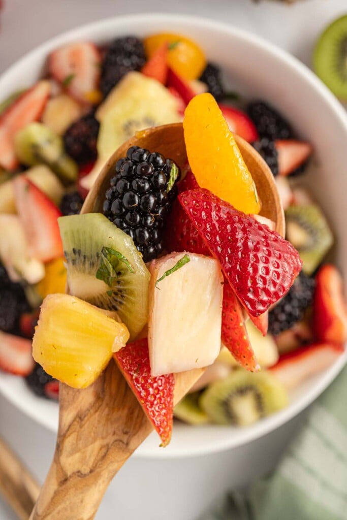 A wooden spoon holding up a scoop of fruit salad with a white bowl of the salad in the background.