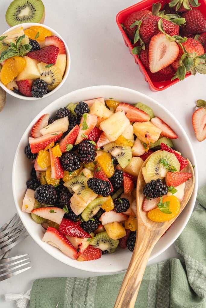 An overhead shot of a white bowl of fruit salad with a wooden spoon in the bowl.