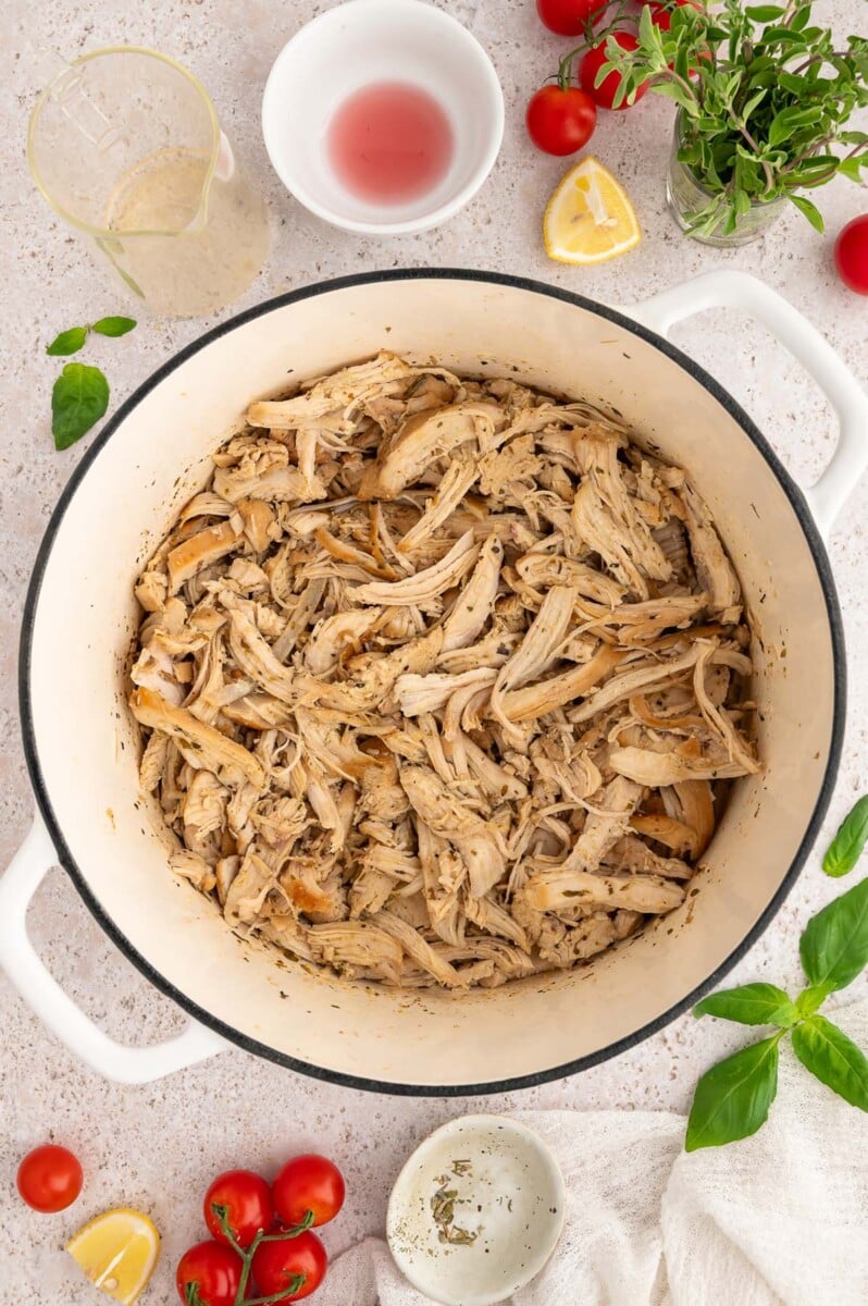 Greek Shredded Chicken in a dutch oven with ingredients on the side.