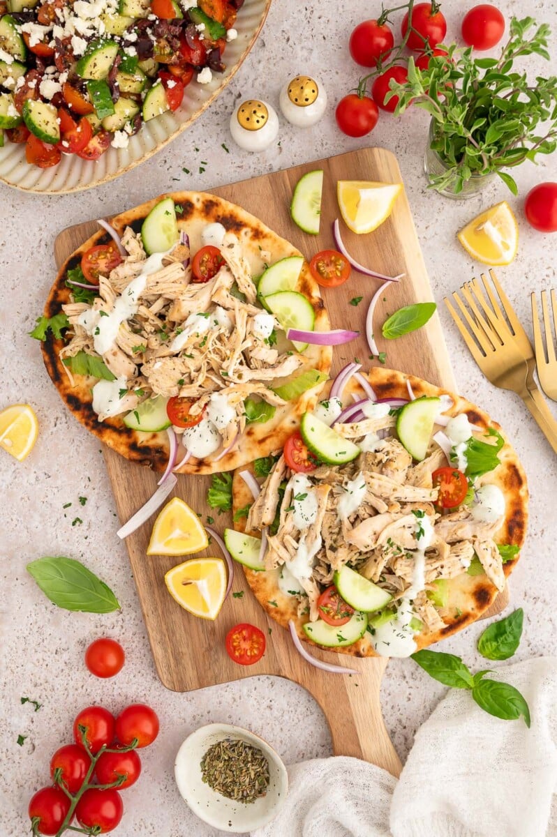 An overhead shot of two greek chicken pitas with cherry tomatoes, sliced cucumbers, and red onions on a wooden board.